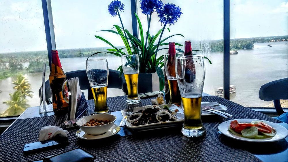 Caption: A photo of a table set with different dishes and several glasses of beer, next to a window overlooking a river in Alappuzha, India. (Local Guide @NanduMuraleedharan)