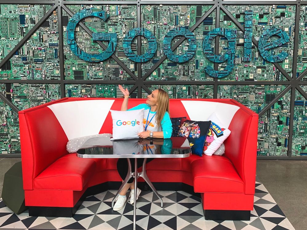 Caption: A photo of Penny sitting on a red sofa holding a pillow and pointing at the letters Google on a wall. (Courtesy of Local Guide @PennyChristie)