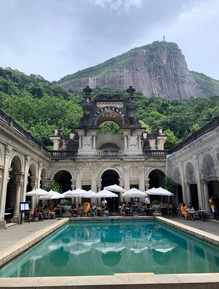 Caption: A photo of the mansion inside Parque Lage, with a pond in front of it and a hill behind it, with the statue of Christ the Redeemer on top.  (Local Guide Juan Ignacio Sepúlveda)