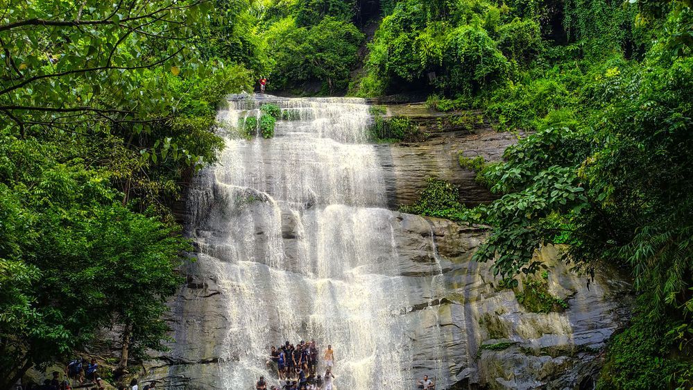 Caption: A photo of the Khoiyachora Waterfall surrounded by green trees and cascading down rocks over some people at its foot. (Local Guide @ShafuilB)