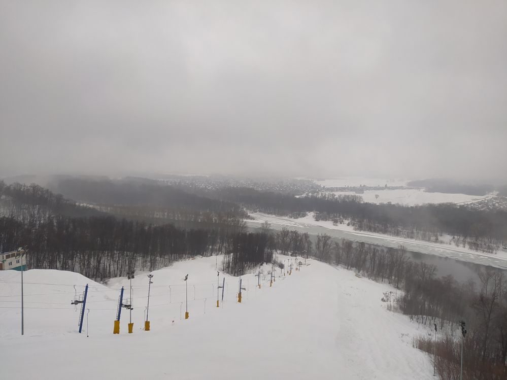 Caption:  A Ski jumping field, toward the left are the cable car support poles. Far in the bottom is river Ufa and surrounding villages.