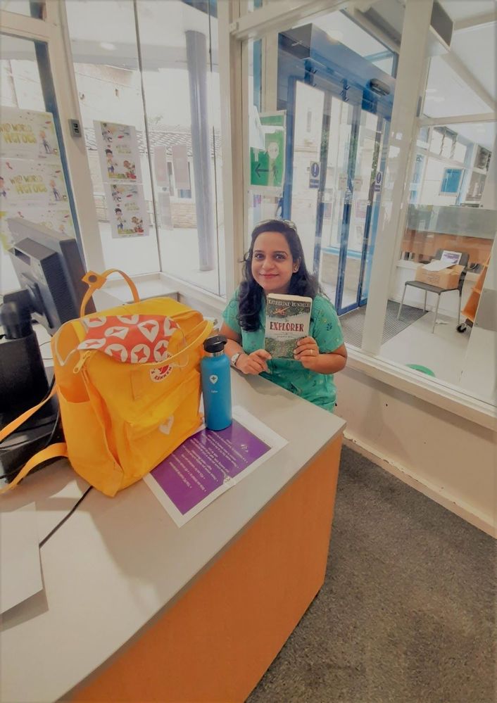 Caption: A photo of Isha holding a book and sitting behind a desk with a Local Guides backpack and a tumbler on it during her meet-up for book lovers. (Courtesy of Local Guide @Globe_trotter_Isha)