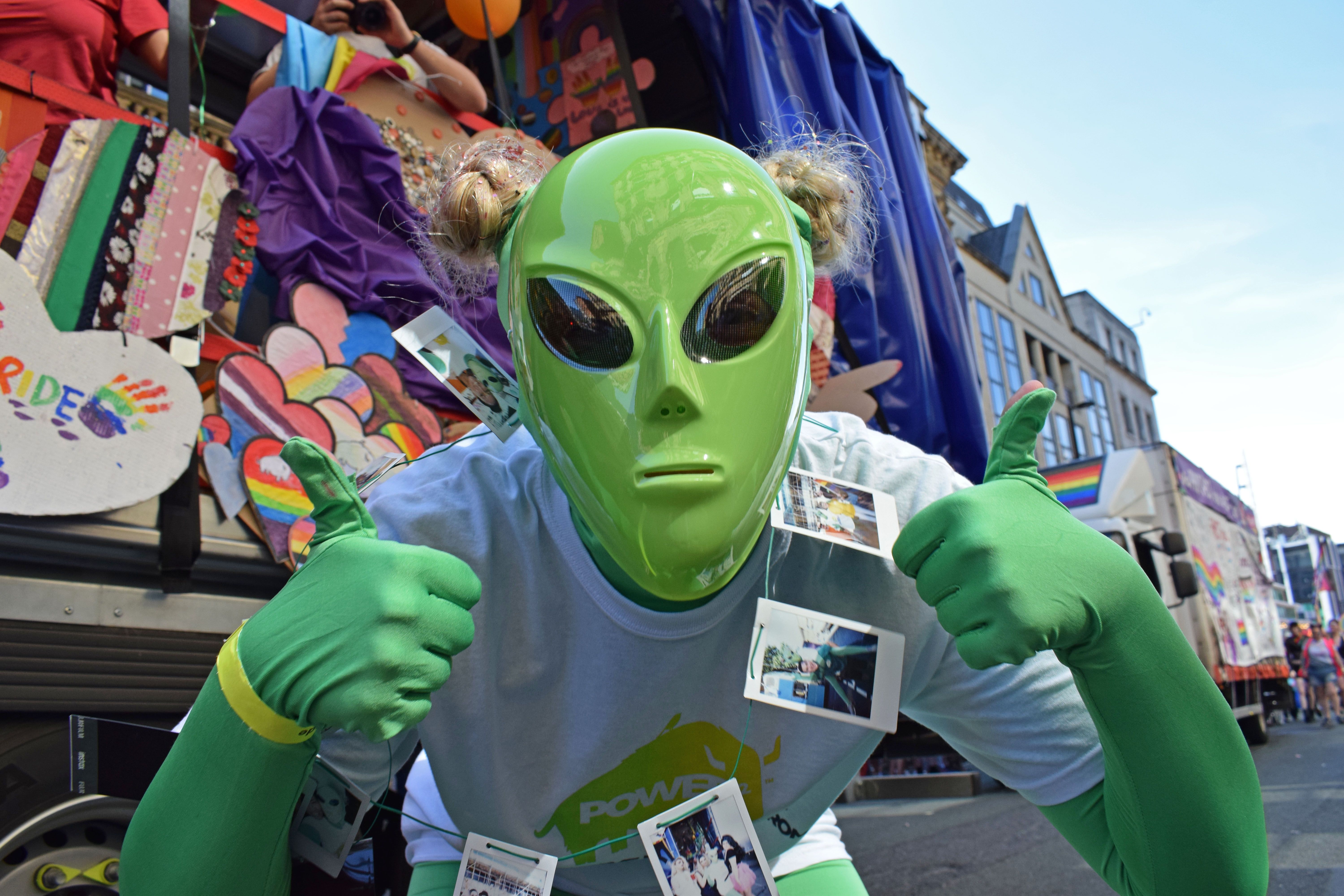 A participant in alien costume with green mask and large eyes making a thumbs up sign