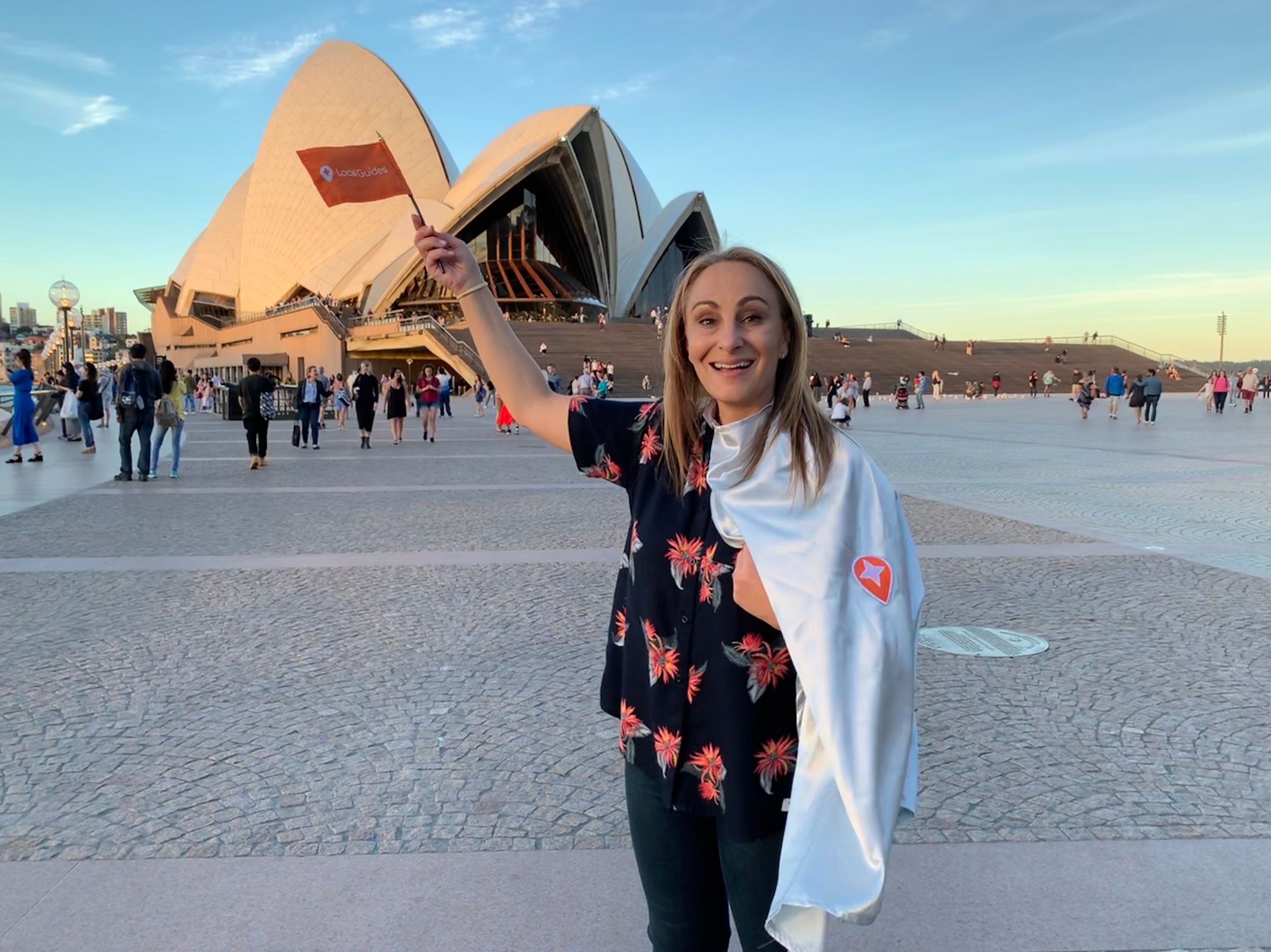 Caption: A photo of Penny holding a Local Guides flag and a cape with the Local Guides pin and posing in front of the Sydney Opera House. (Courtesy of Local Guide @PennyChristie)