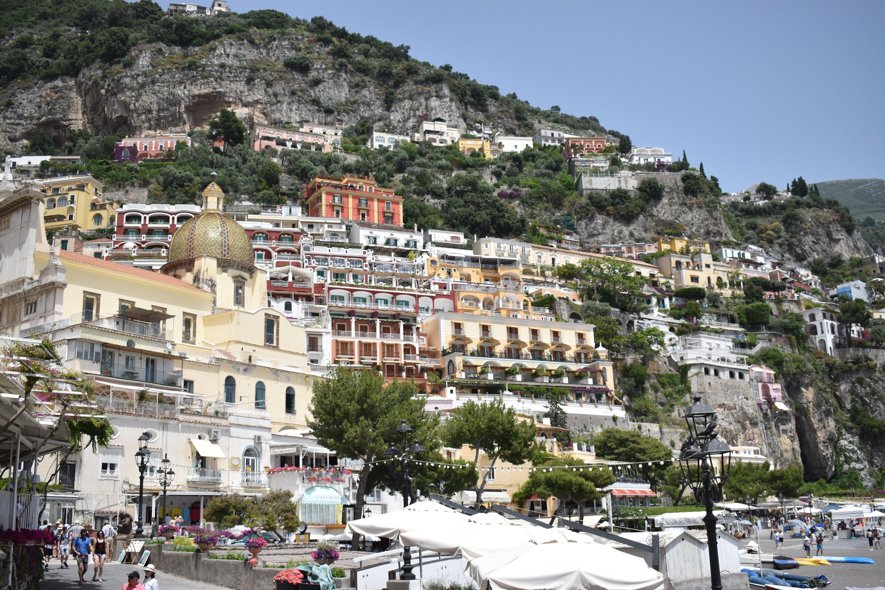 Caption: A photo of the colorful, terraced houses close to the beach in Positano and the rocky hills above them. (Local Guide @MoniDi)