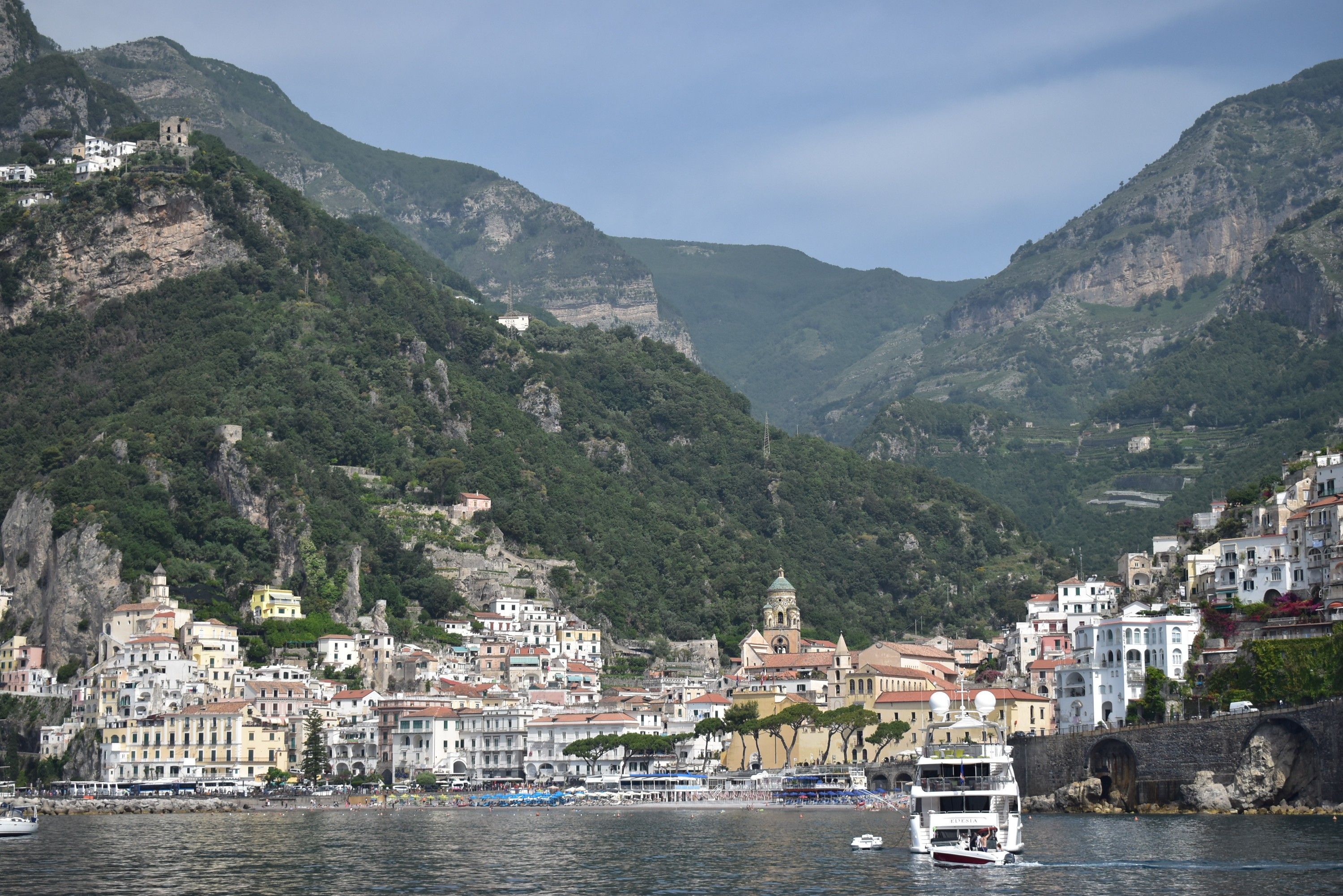 Caption: A photo from a boat of Amalfi, nestled between the mountains, with a yacht floating on the water in its bay. (Local Guide @MoniDi)