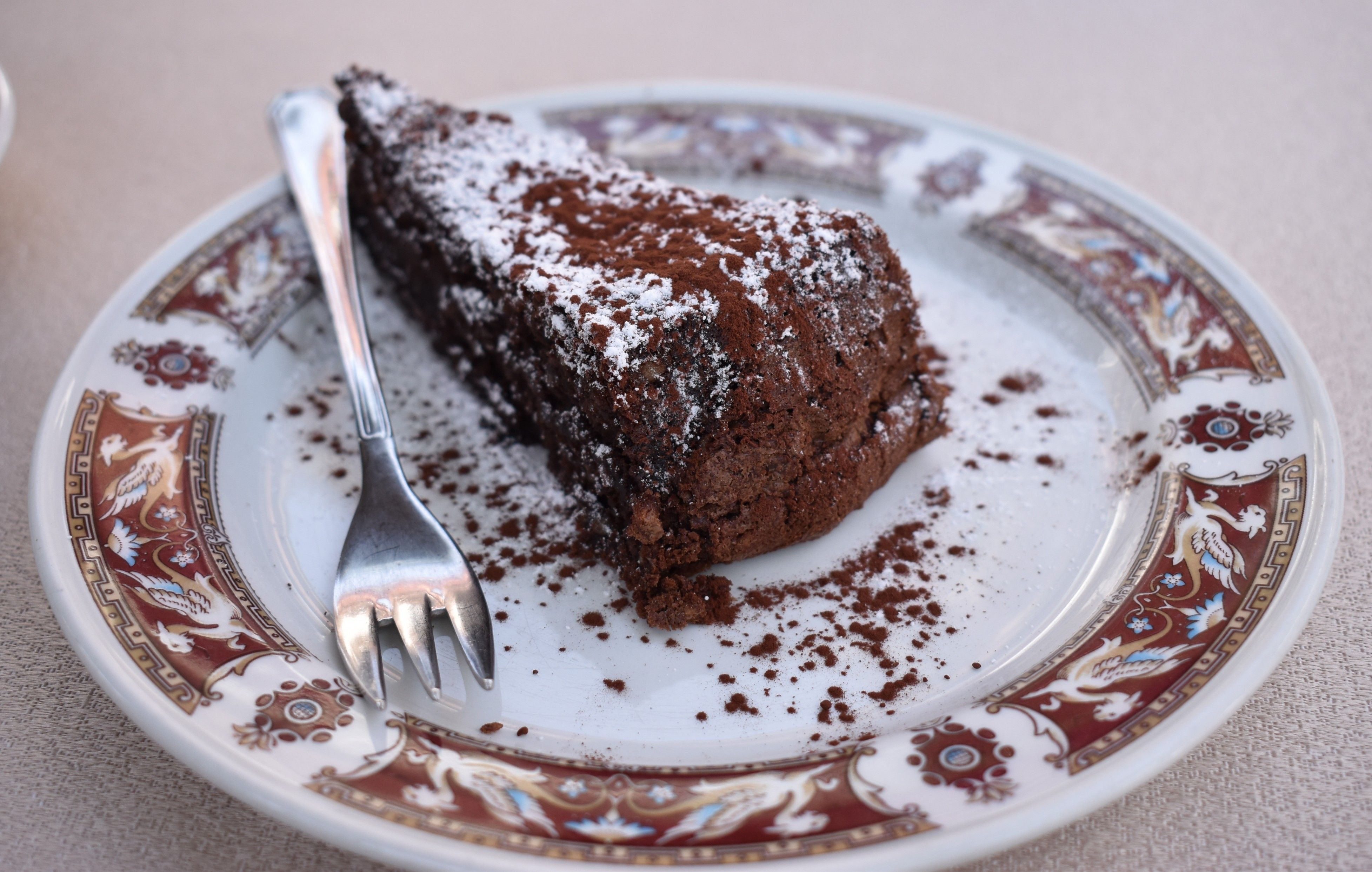 Caption: A photo of a piece of chocolate Torta Caprese cake, sprinkled with powdered sugar and cocoa, and served on a patterned plate with a fork on the side. (Local Guide @MoniDi)