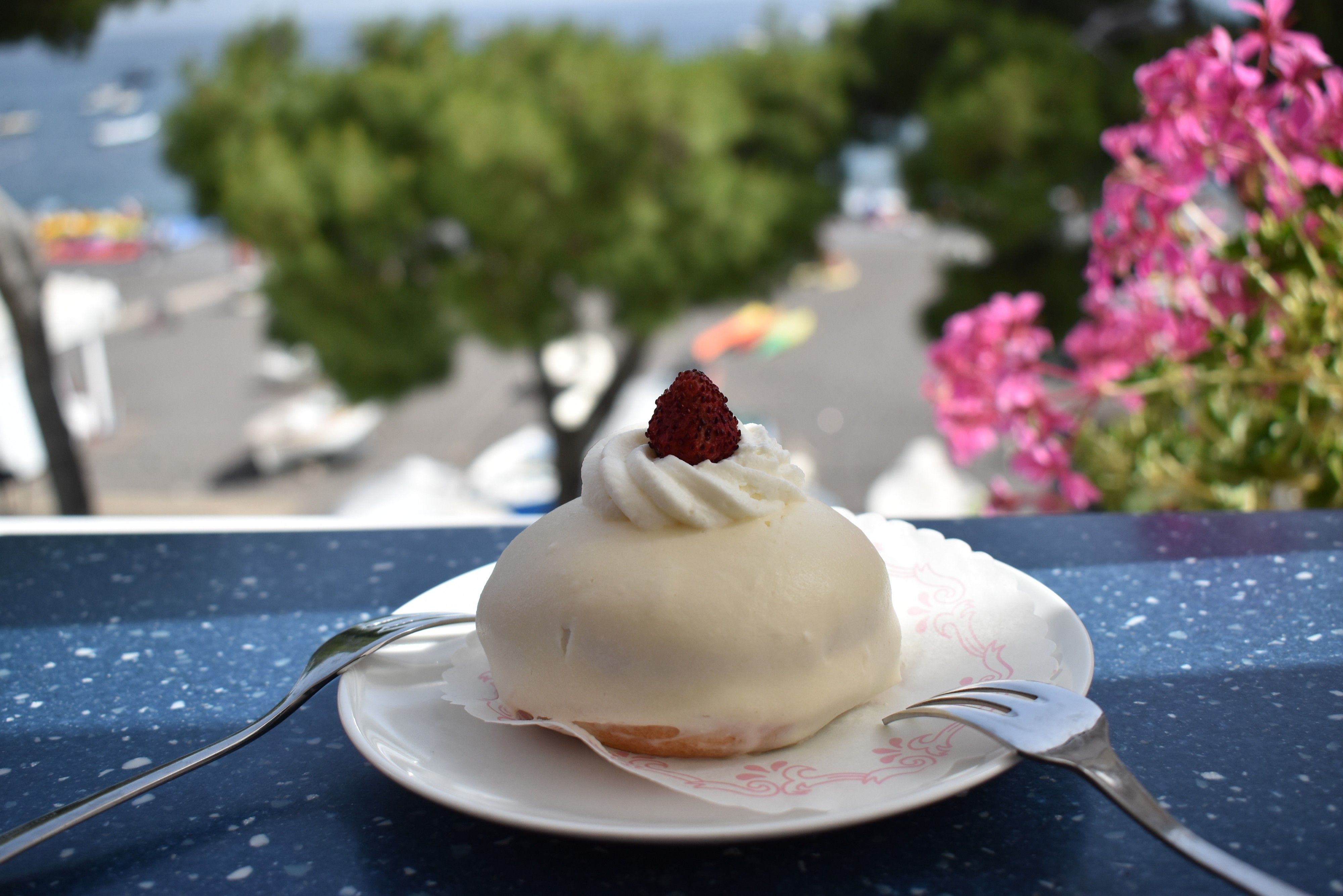 Caption: A photo of a small white round Delizia al Limone cake with whipped cream and a wild strawberry on top and two forks placed on the plate. Pink flowers and the Positano beach are visible in the background. (Local Guide @MoniDi)