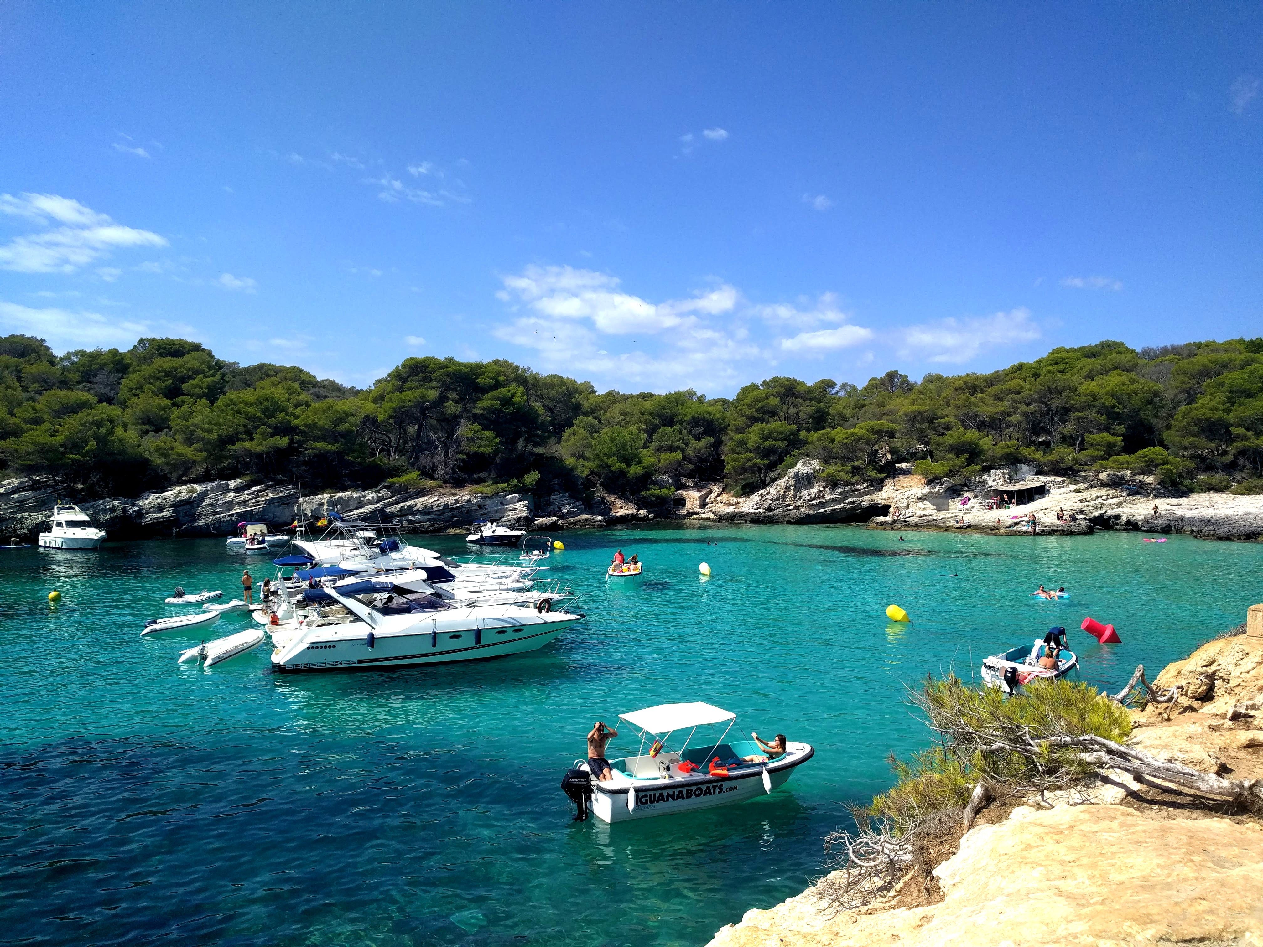 Caption: A photo of several boats floating on the turquoise waters near a rocky beach in Menorca, Spain. (Local Guide @MoniDi)