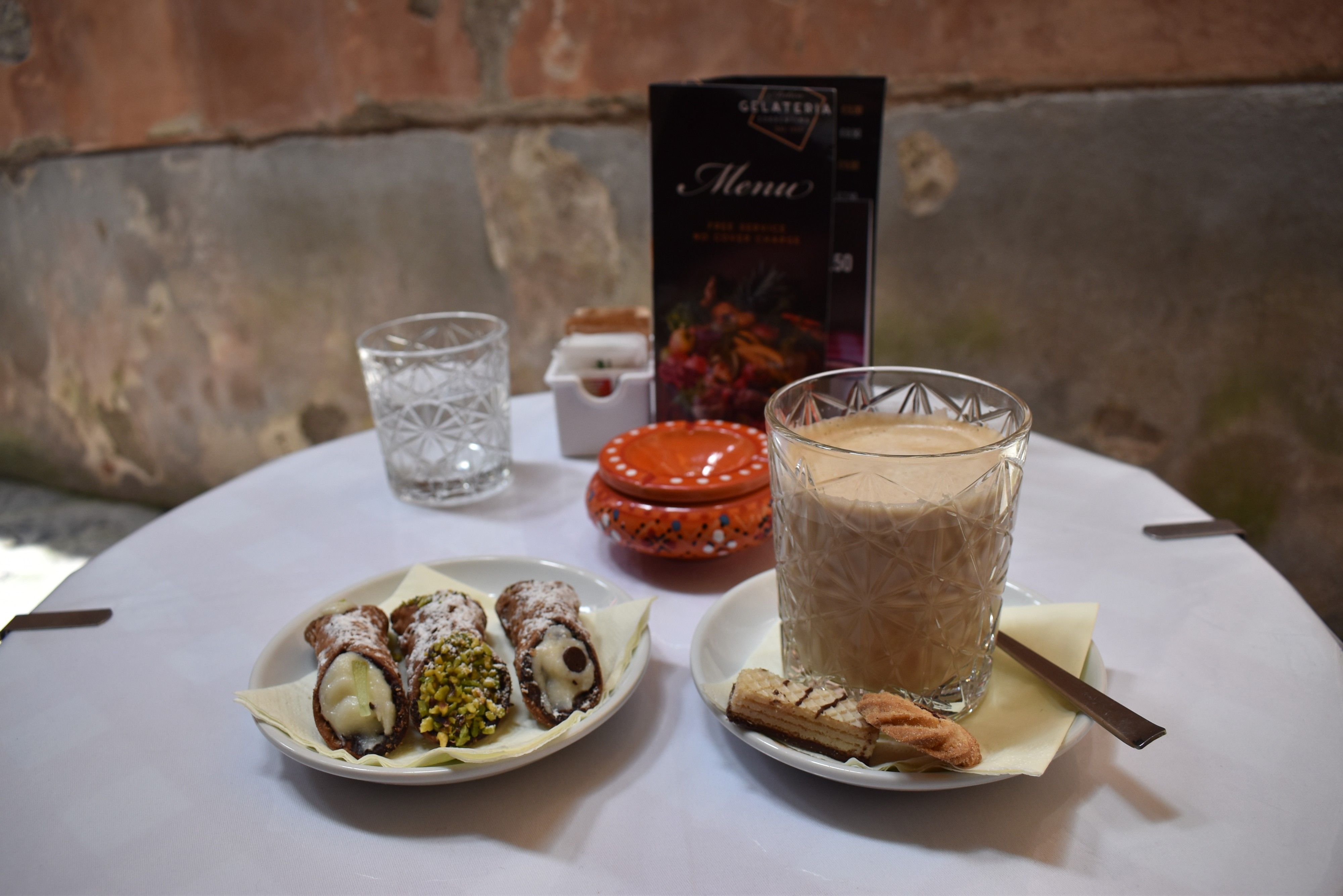 Caption: A photo of three Italian cannoli pastries on a plate and a cup of caffe latte served with biscuits on the side, arranged on a table outside a café. (Local Guide @MoniDi)