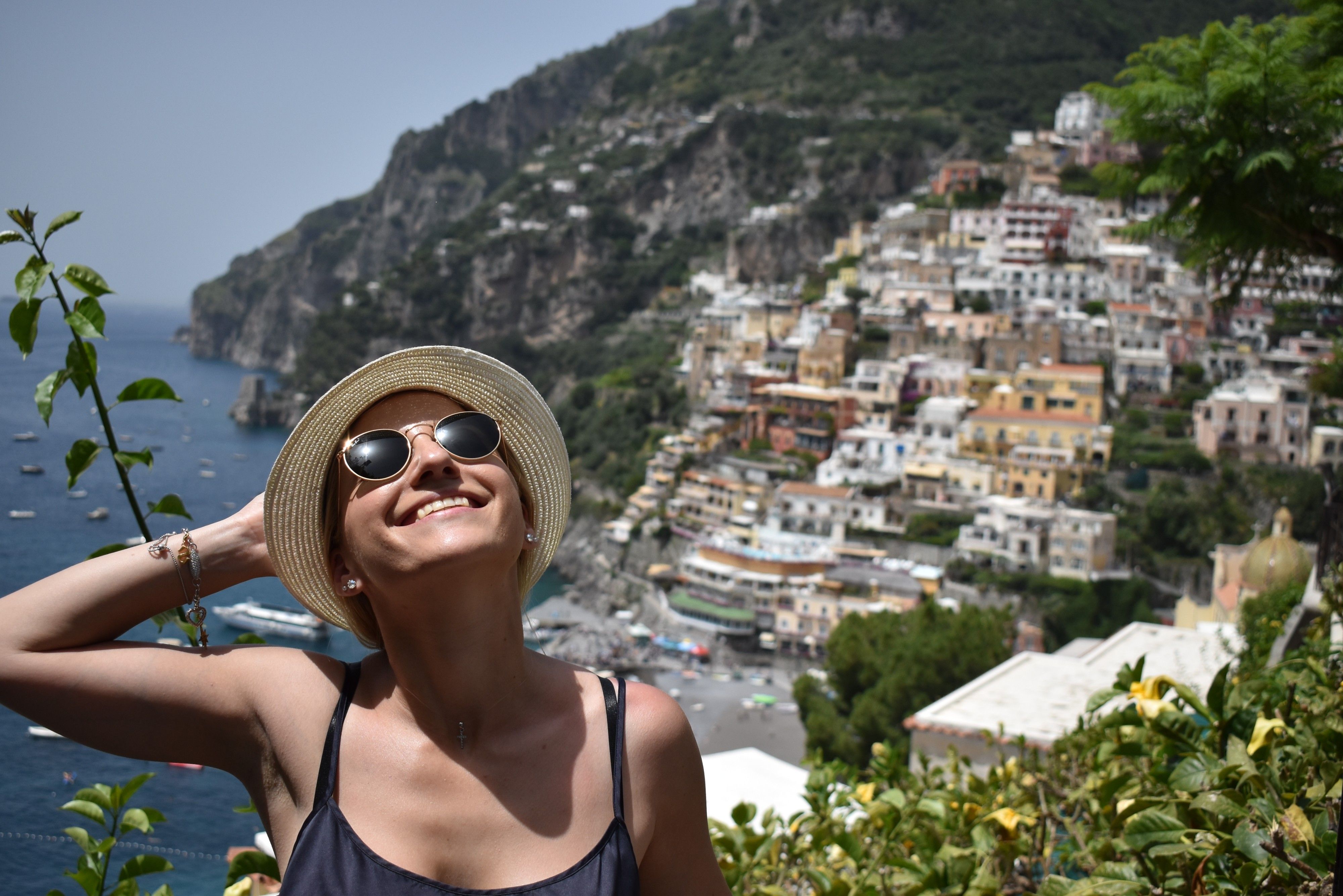 Caption: A photo of Google Moderator @MoniDi with sunglasses and a straw hat, looking up at the sun and smiling, standing on a terrace overlooking Positano, Italy. (Local Guide @MoniDi)