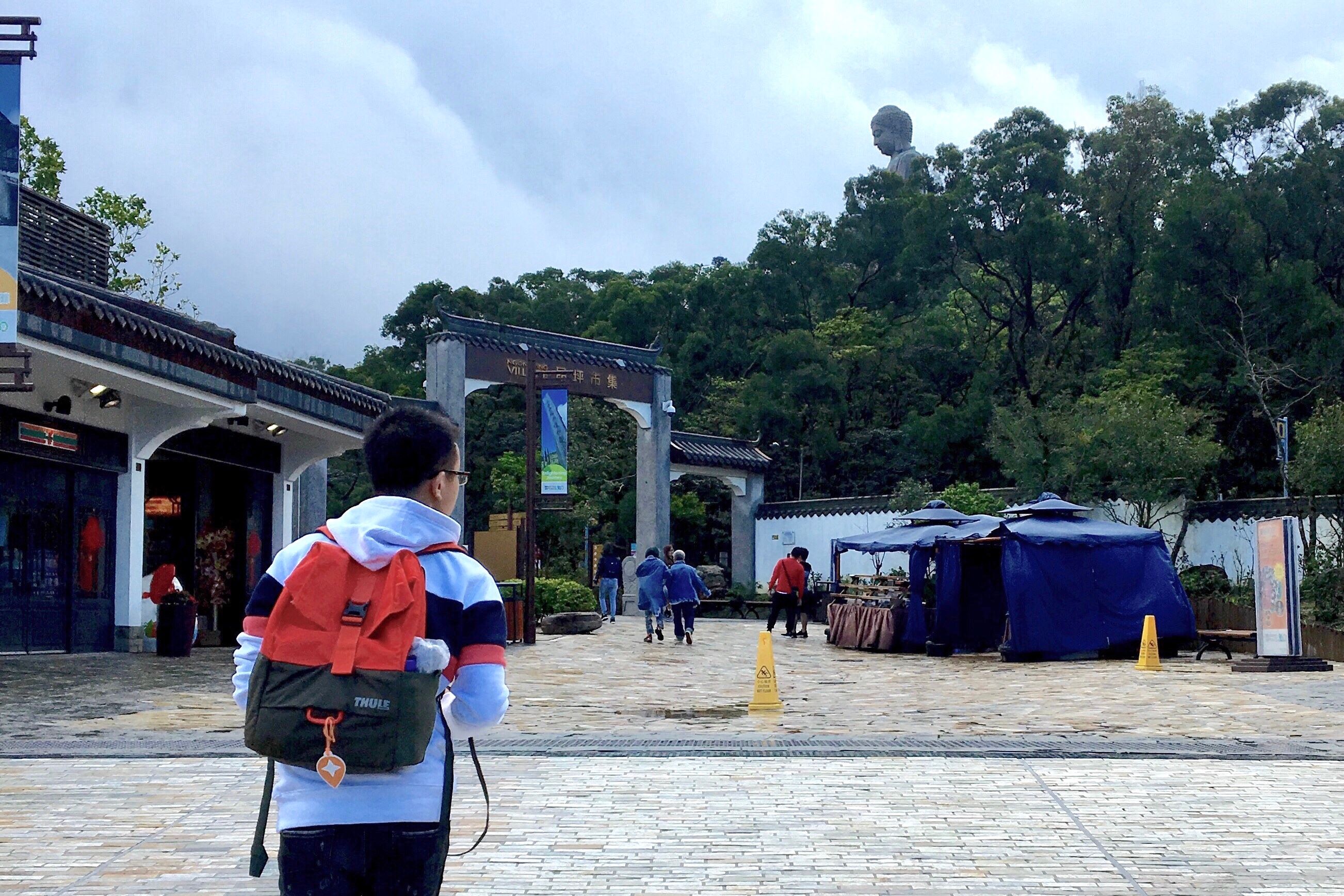 Photo of myself carrying a bag with a small Local Guide name tag  attached to my backpack and walking towards  the Tian Tan Buddha Statue in Hong Kong.