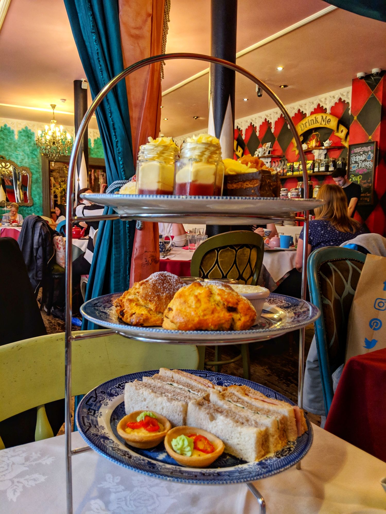 Afternoon tea on a cake stand. Sandwiches on the bottom layer, scones cream and jam on the middle layer and  cakes on the top layer. In the background is the serving area with eat me and drink me in large writing on the wall