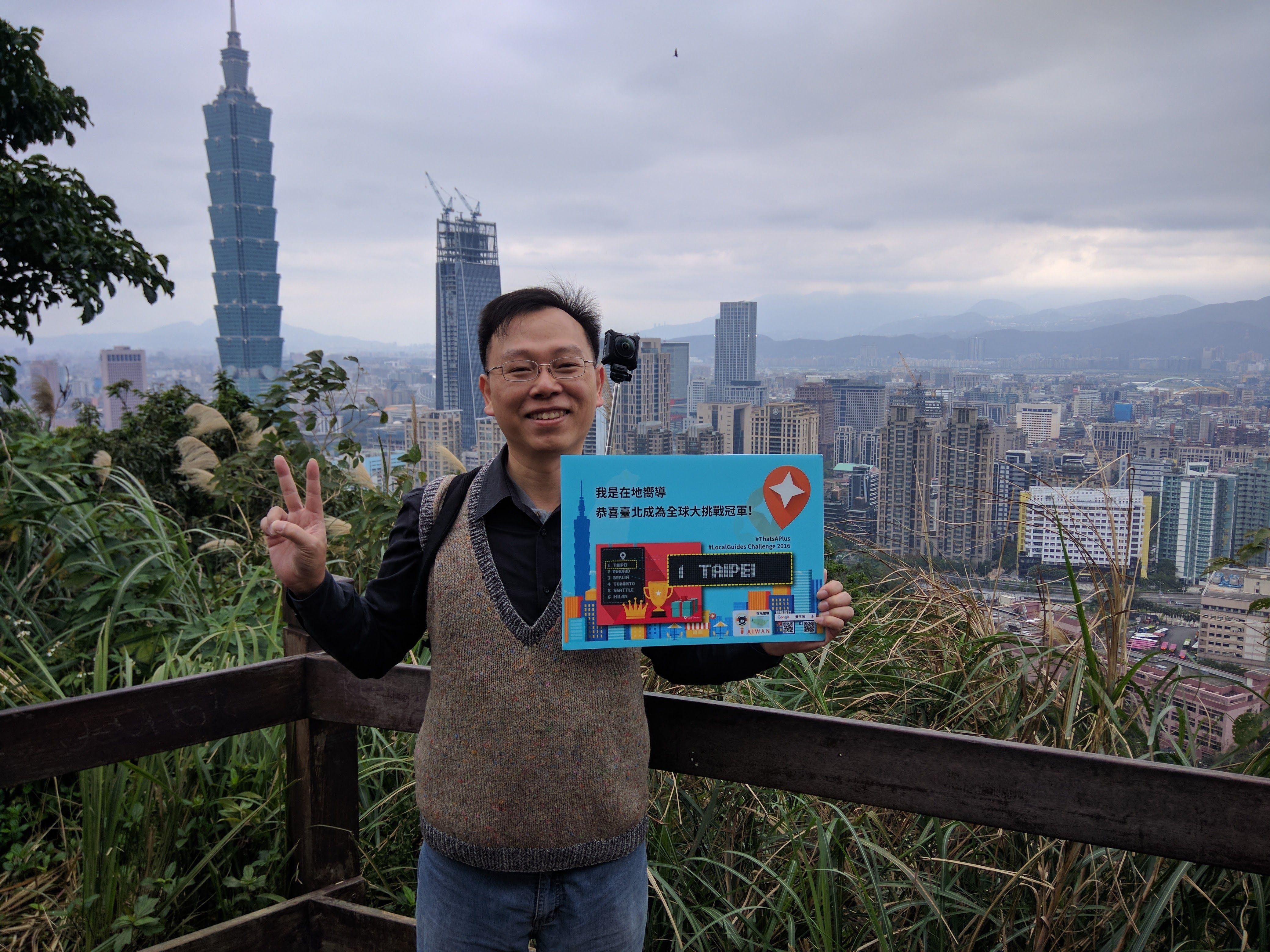 Caption: A photo of Sampson on the Xiangshan Hiking Trail in January 2017. The sign celebrates Taipei’s victory in the LGChallenge 2016. The tallest building in Taiwan, the Taipei 101 Tower, is behind him. (Local Guide @SampsonF)