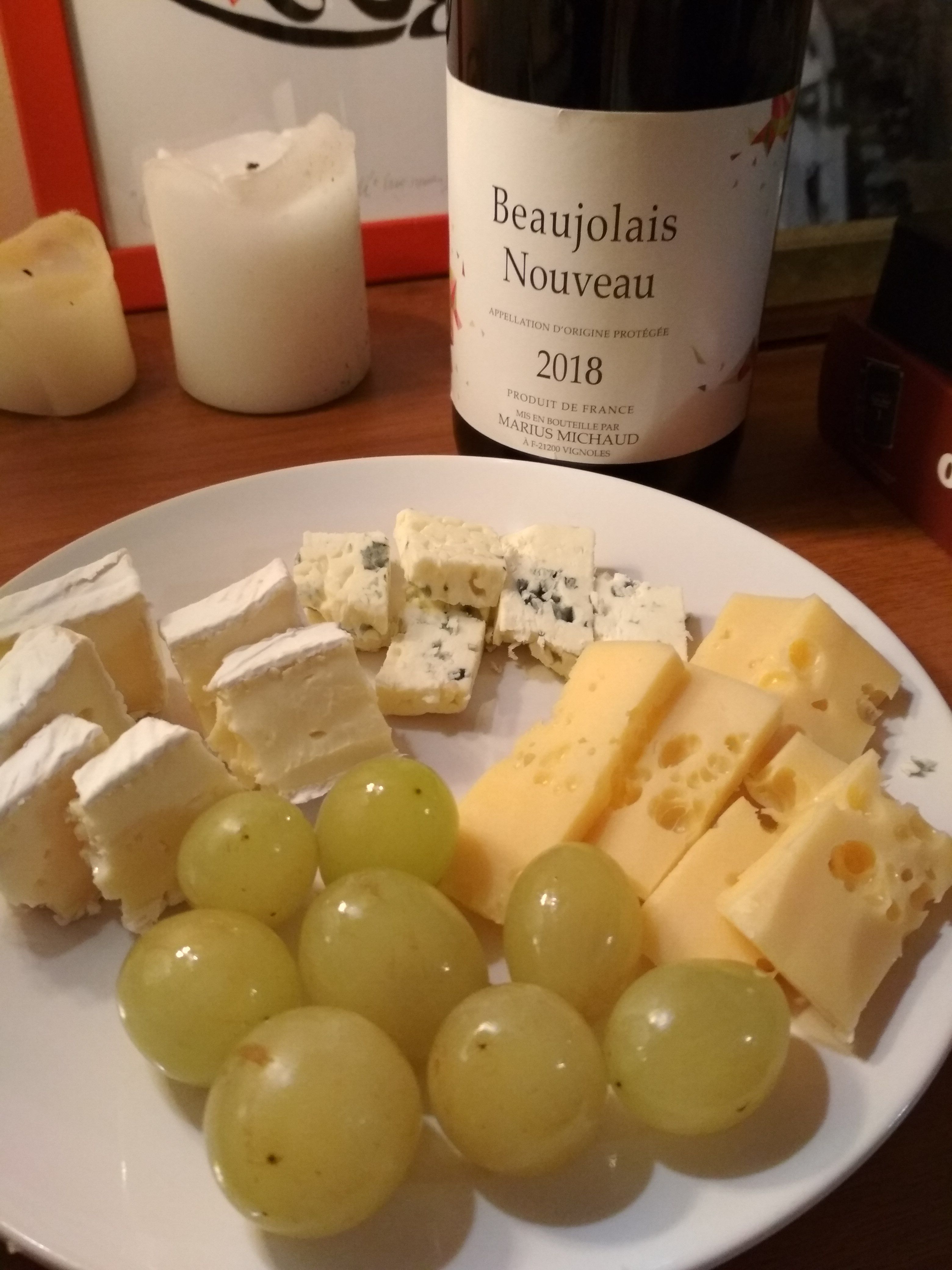 Caption: A photo of a plate with grapes, brie cheese, blue cheese, and Swiss cheese. There are two candles, and a bottle of red Beaujolais Nouveau wine in the background. (Local Guide @DeniGu)