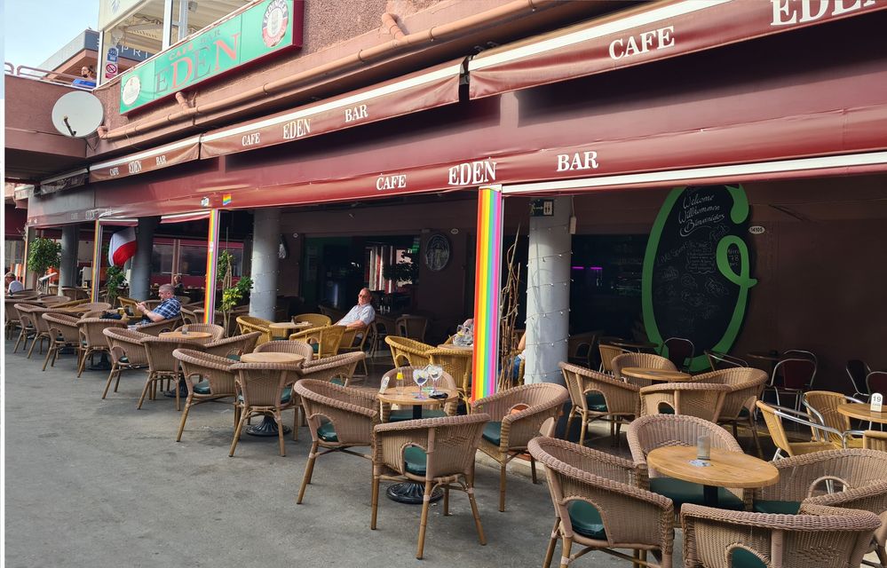 Caption: A photo of the outdoor seating area of the Cafe Bar Eden in Maspalomas, Spain, with a rainbow flag in the background. (Local Guide @HeyitsNicho)