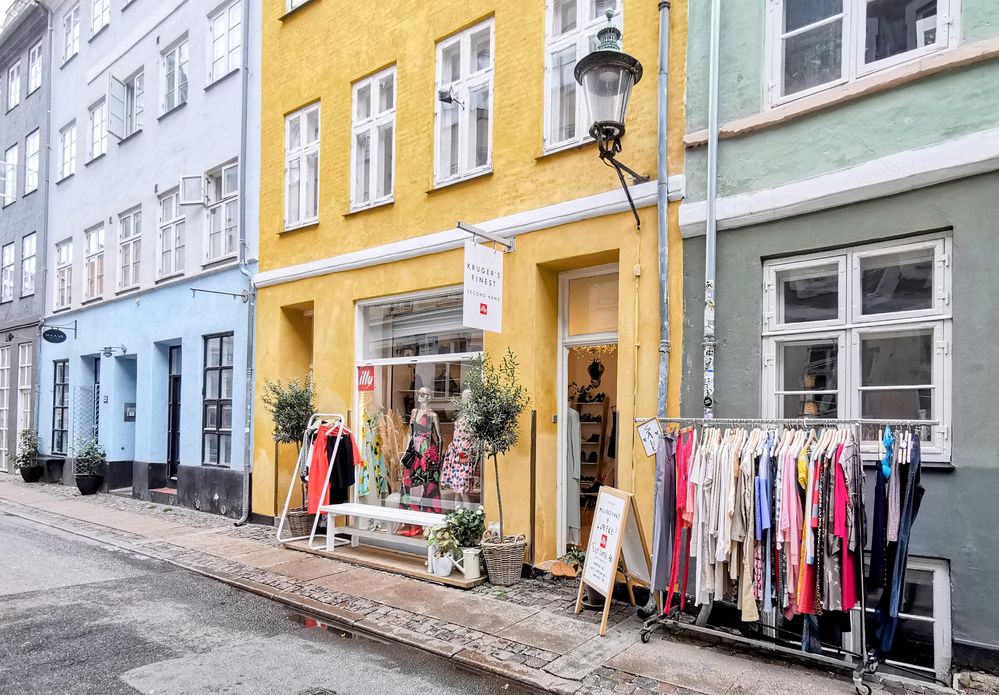 Caption: A photo of blue, yellow, and green storefronts with clothes racks, a bench, and plants in front of the yellow store. (Local Guide @MortenCopenhagen)