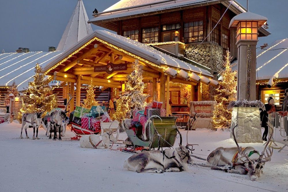 Caption: An exterior photo of a snowy wood chalet with Christmas trees decorated in white lights, sleighs filled with presents, and reindeer at Santa Claus Village in Rovaniemi, Finland. (Local Guide Mauro Scardoni)