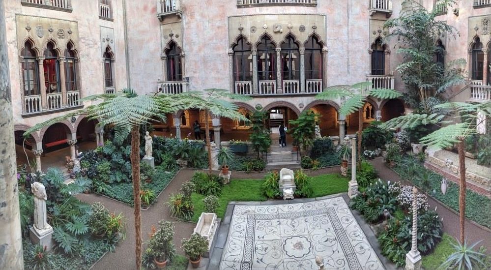 My photo of the courtyard of the Isabella Gardener Museum. It shows a tiled center area, palm trees and other lush plants, various pieces of sculpture and the façade opposite where I was standing. The courtyard is 3 stories high with a glass roof