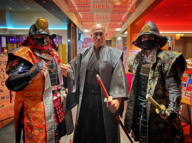 Caption: A photo of Masa in traditional Japanese attire holding a sword and flanked by two people in samurai warrior clothes.