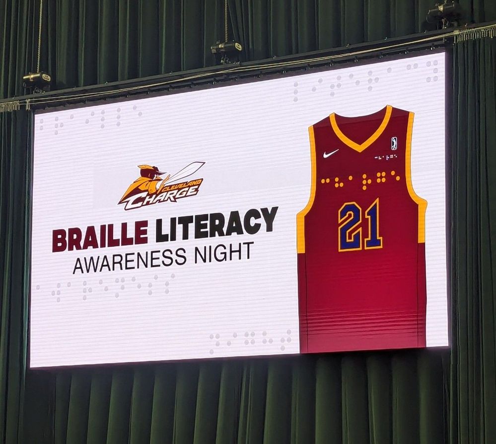 Caption: a photo of the large LED screen displays the braille embroidered jerseys, in promotion of Braille Awareness Night