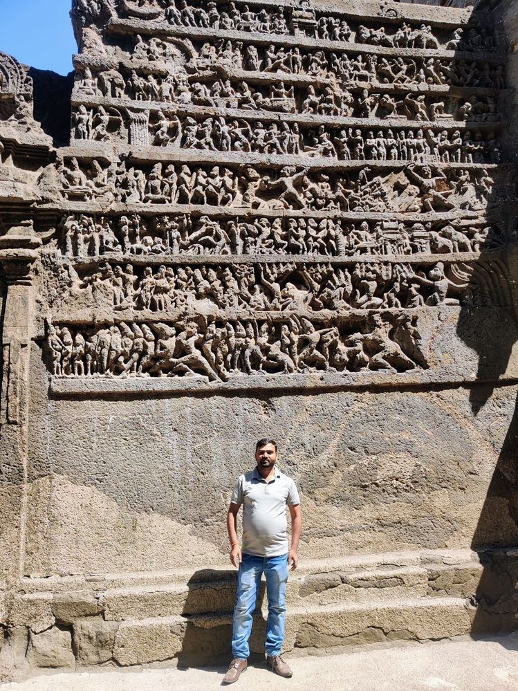Photograph of LG NandKK before a temple wall full of rock-cut statues of hindu dieties dancing and battling