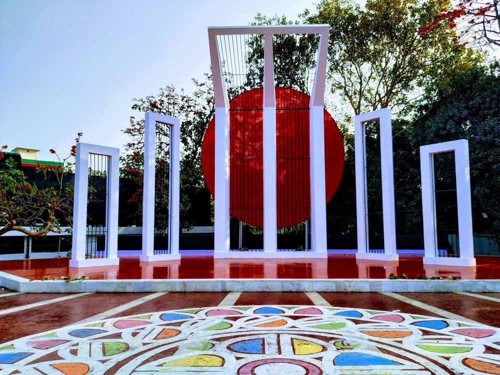 Caption: A photo of the Central Shaheed Minar monument in Dhaka, which consists of five pillars with a big red circle behind the central pillar, and a circular drawing in white, blue, pink, green and orange on the ground in front of it. (Courtesy of Local Guide @Papel_Mahammud)