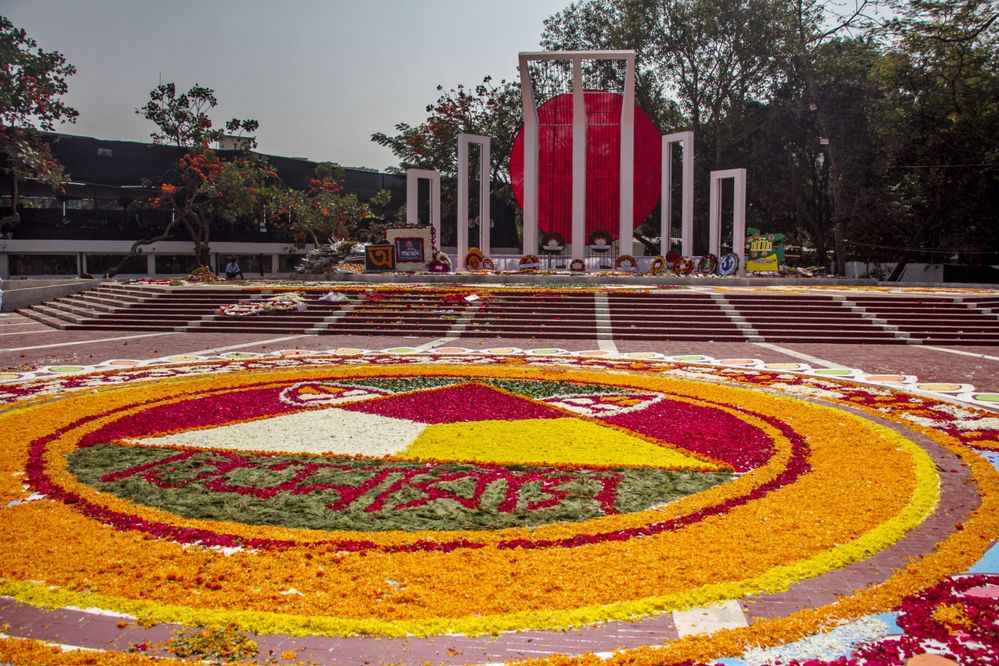 Caption: A photo of the Central Shaheed Minar monument in Dhaka, with wreaths placed against the pillars and a large circular design made out of red, yellow, orange, white, and green petals on the ground in front of it. (Courtesy of Local Guide @MohammadPalash)