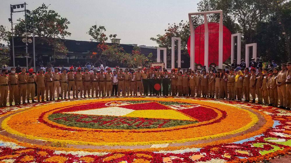 Caption: A photo of a platoon standing around a large circular design made out of red, yellow, orange, white, and green petals on the ground in front of the Central Shaheed Minar monument in Dhaka. (Courtesy of Local Guide @MohammadPalash)