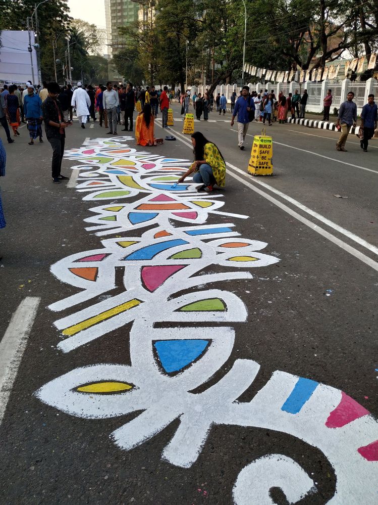 Caption: A photo of a person drawing a vibrant design of white, blue, pink, yellow, green, and orange on a street in Dhaka, with many other people walking around. (Courtesy of Local Guide @Papel_Mahammud)
