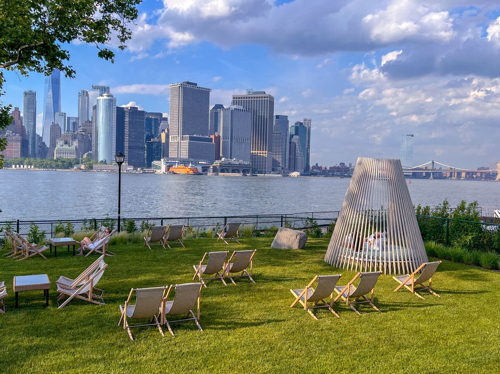Caption: A photo of deck chairs, tables, and an outdoor daybed on a lawn at QC Spa, one of the most relaxing spots in New York City, with the city’s skyline in the background. (Courtesy of Local Guide @Kiariladyboss)