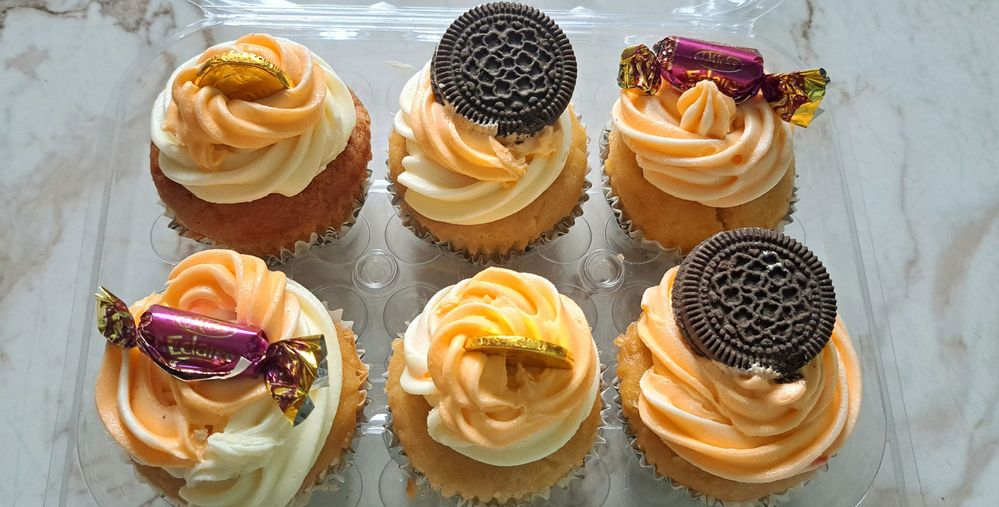 Caption: A photo of the pack of six cupcakes I ordered from a woman owned buisness in my area. A confectionery shop close to me. Note the orange and white Icing to suit connect. Note also the biscuits sweets and etc used to dress the cupcakes. @SholaIB