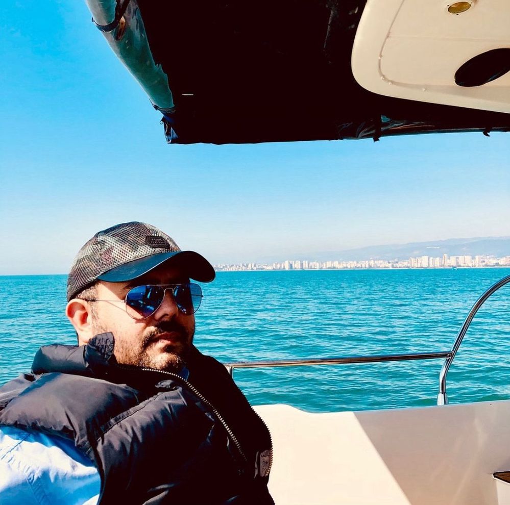 Caption: A photo of Local Guide Burak on a yacht at the Mersin Marina in Turkey, with the sea and Mersin city skyline behind him. (Courtesy of Local Guide @Gezendunyali)
