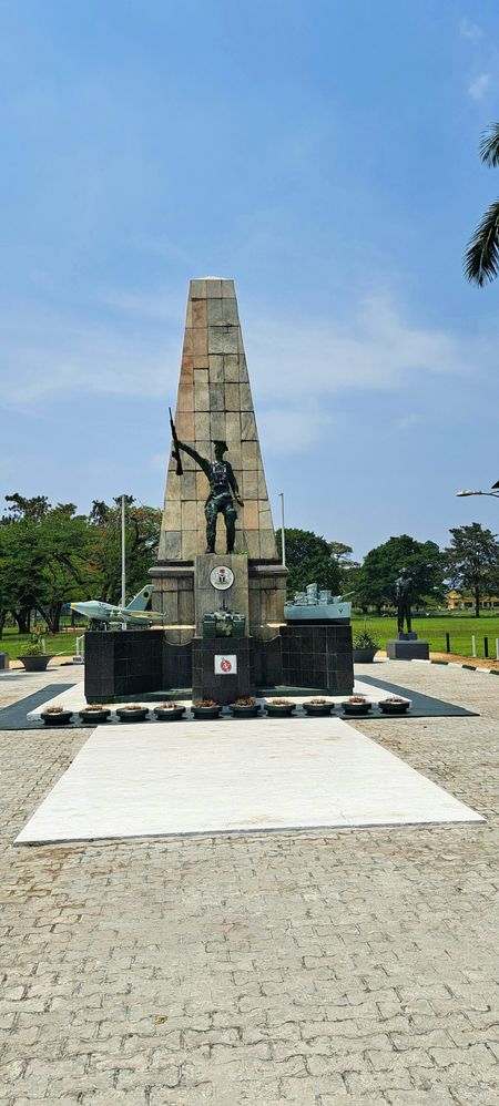 Caption: A photo of the whole statue of the monument itself against  which constitute of a  soldier standing with his gun An aircraft,A ship and an armoured tank. The Nigerian coat of arm as well. All the image against the green lawn and blue sky in the background. @SholaIB