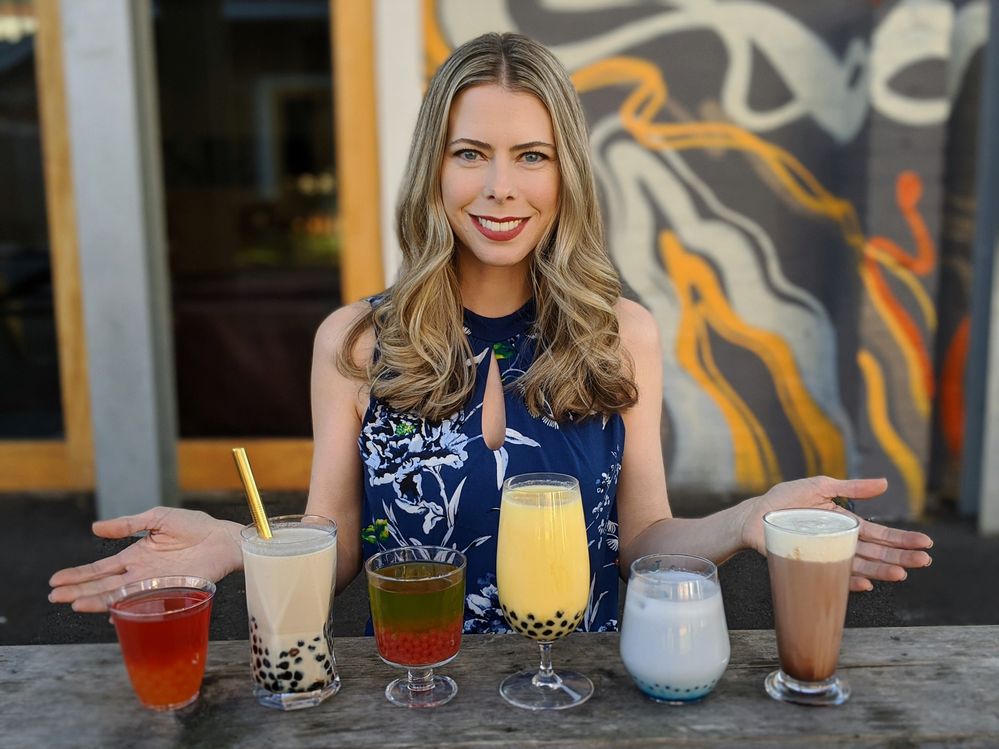 Caption: A photo of Kristin smiling, with six glasses of different bubble tea flavors on the table in front of her. (Courtesy of Local Guide @TheRealKristin)