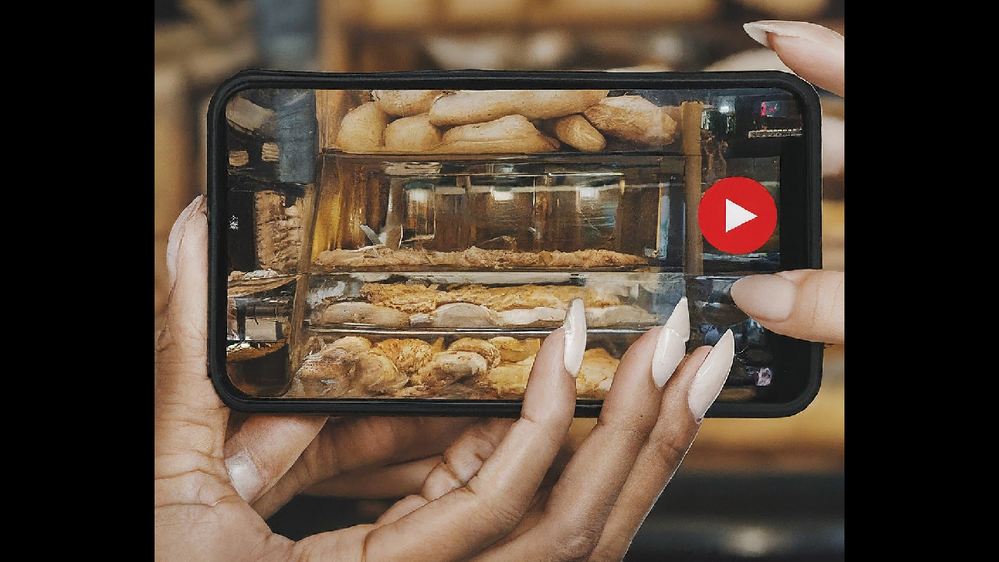 Caption: Image of a lady using her smartphone and taking a video in a bakery shop.