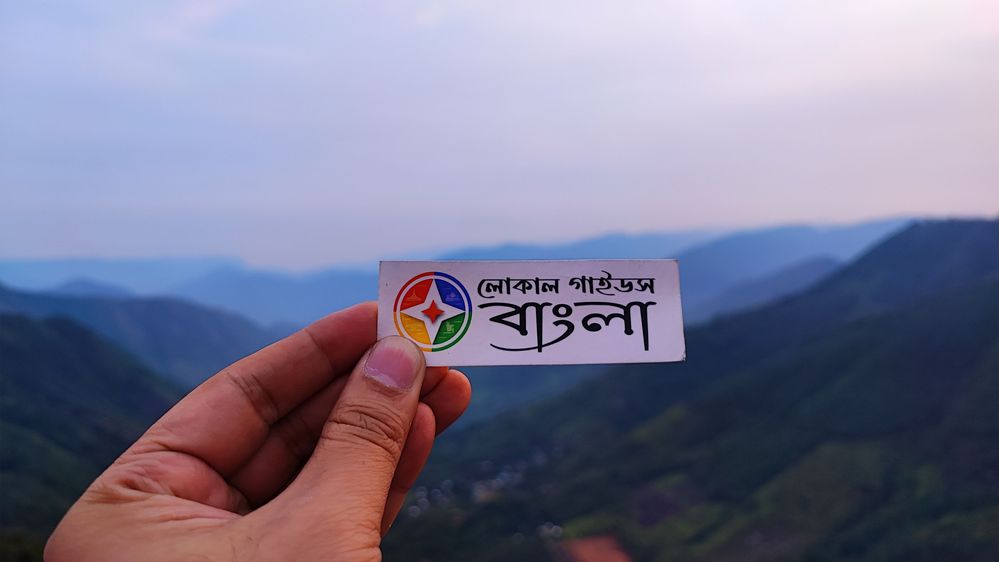 A photo of a hand holding a sticker of Local Guides Bangla and showing a beautiful view of Meghalaya