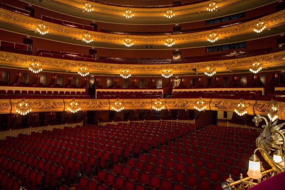 Caption: A photo by Carlos Pareja of the grand Liceu Opera Barcelona in Spain featuring the empty theater with red seats, tiers of golden balconies, and chandeliers.