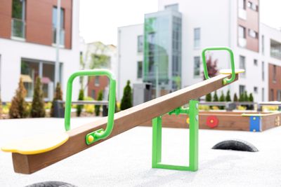 A wooden seesaw with green handlebars and yellow seats in the foreground and other playground equipment and surrounding buildings in the background (Getty Images)