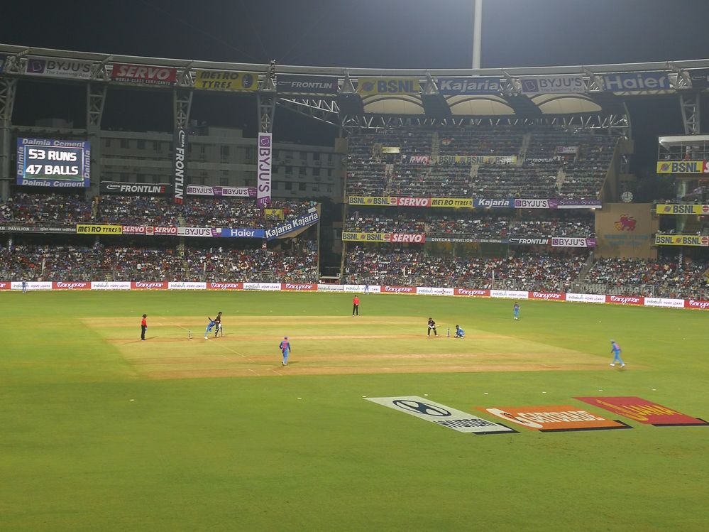 Caption: A stadium filled with people watching an ongoing cricket game, photo by Neil Shah