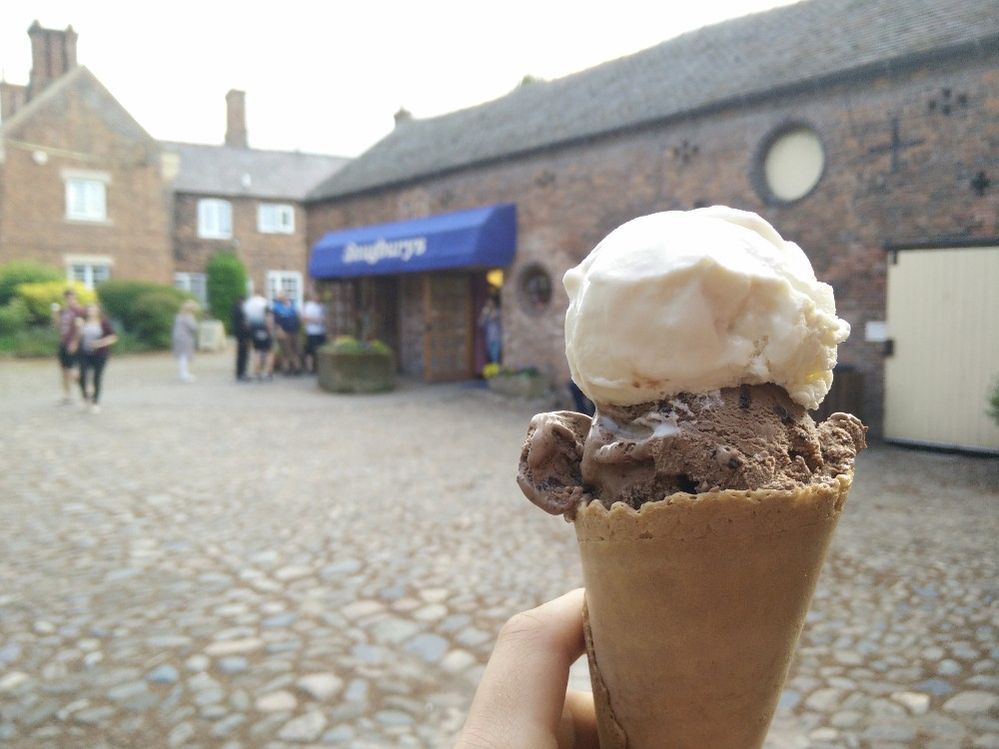 Caption: A photo of someone holding an ice cream cone with two scoops in front of the cobblestone courtyard and bright blue awning of Snugburys Ice Cream in Nantwich, England. (Local Guide Michael Hunter)