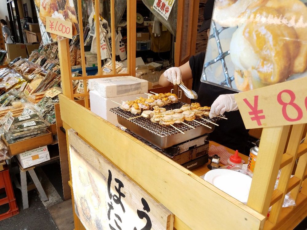 Caption: A photo of a stall at Tsukiji Market in Tokyo, Japan where a man is roasting fish and shellfish on skewers on a small tabletop grill. (Local Guide Kazuhisa SHIHOMMATSU)