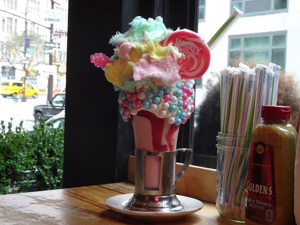 Caption: A photo of Black Tap’s “Cotton Candy Crazy Shake” that features a pink milkshake in a glass decorated with icing and round candies, as well as pink and blue cotton candy, a pink and white lollipop, and pink rock candy on top. (Local Guide Kenichi Shimada)