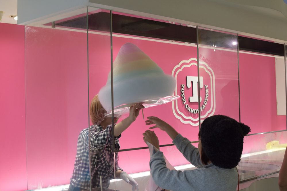 Caption: A photo of a Totti Candy Factory employee handing a customer a large cone of multi-colored cotton candy served on a stick with the shop’s bright pink walls in the background. (Local Guide サニーテディ)