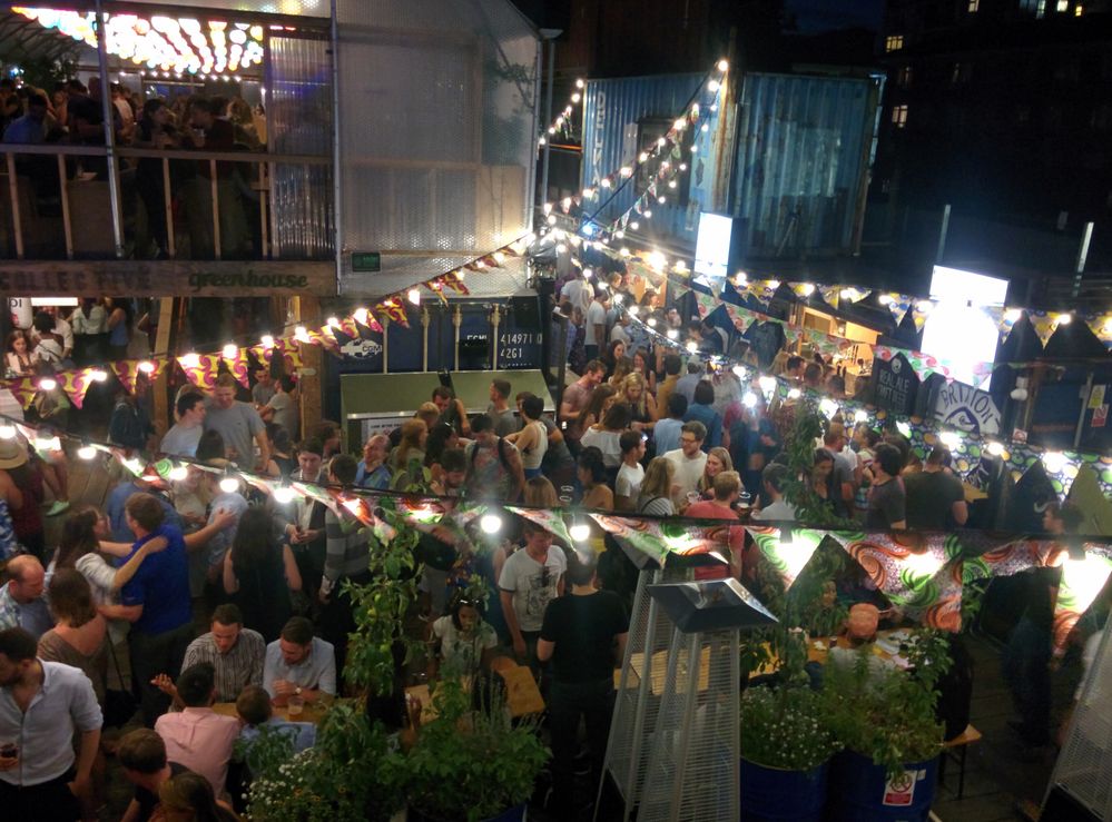 Caption: A photo of a crowd of people dancing and enjoying drinks outdoors at Pop Brixton in London underneath string lights. (Local Guide Meeten Shah)