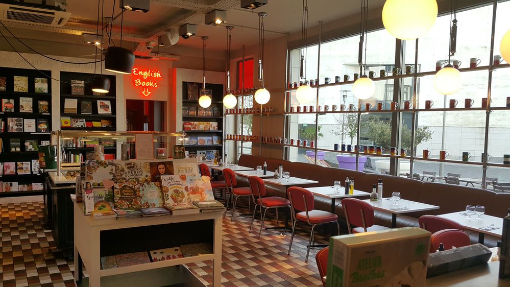 Caption: A photo of the interior of Cook & Book, a bookstore and eatery in Brussels, showing a cookbook display set up on a table and rows of tables and chairs set against large windows. (Local Guide Lahaise Deborah)