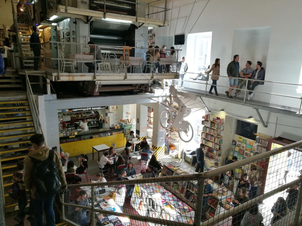 Caption: A photo of Ler Devagar, a bookstore in Lisbon, showing the shop’s multi-level interior with rows of bookshelves, a suspended piece of art of a person riding a bicycle, and a cafe with tables full of patrons. (Local Guide Nuno Ribeiro)