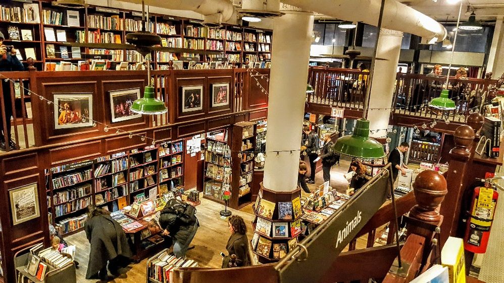 Caption: A photo of Housing Works Bookstore Cafe in New York City, New York, a multi-story, open-concept shop, featuring rows of dark wooden bookshelves and tables stacked with books as well as patrons sitting at tables. (Local Guide Rajiv Dewan)