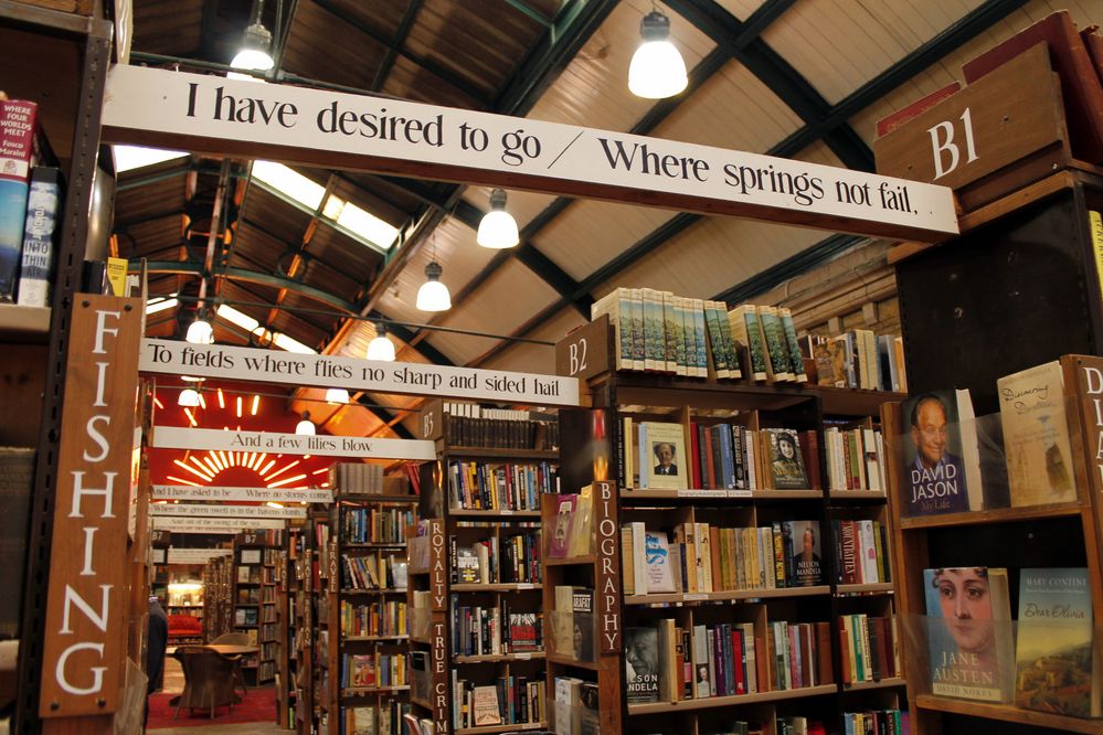 Caption: A photo of Barter Books in Alnwick, England showing rows of bookcases and decorative wood panels hanging from the ceiling with lines from Gerard Manley Hopkins’s poem “Heaven—Haven.” (Local Guide Dave Chick)