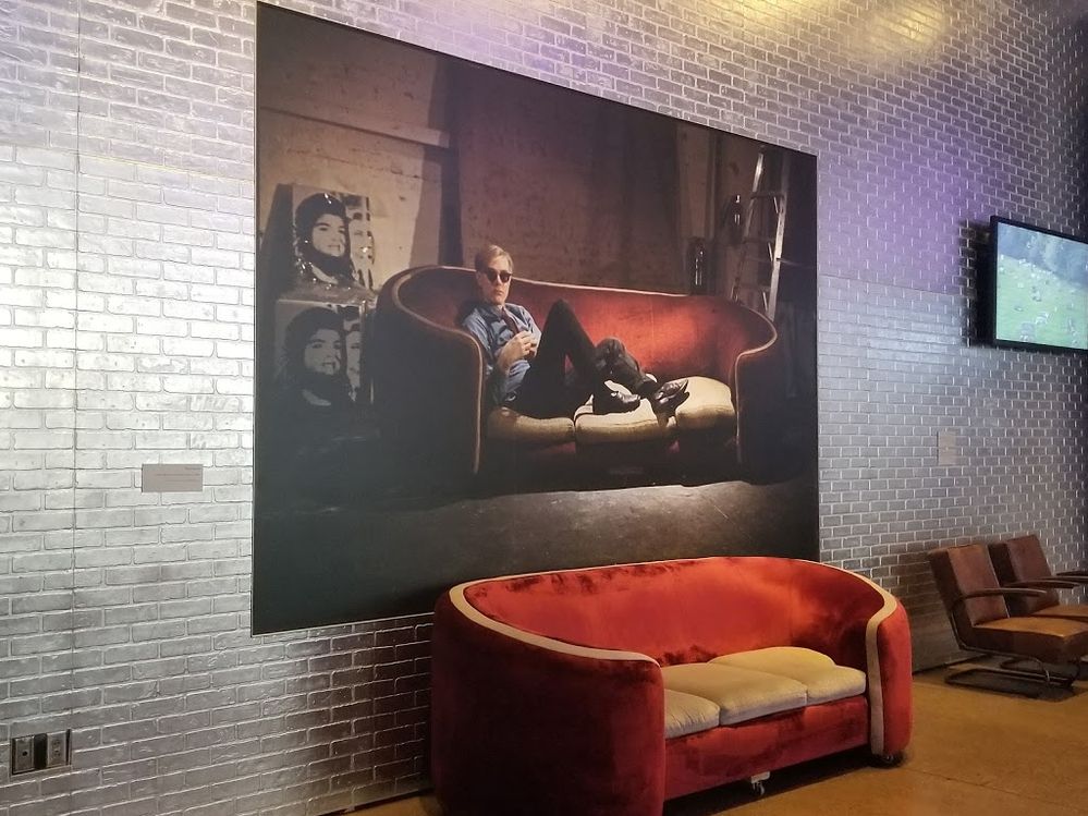 Caption: A photo of The Andy Warhol Museum featuring a large photo of Andy Warhol sitting on a couch hung on a silver brick wall with a similar-looking couch to the one in the photo in front of it. (Local Guide Emily Webb)